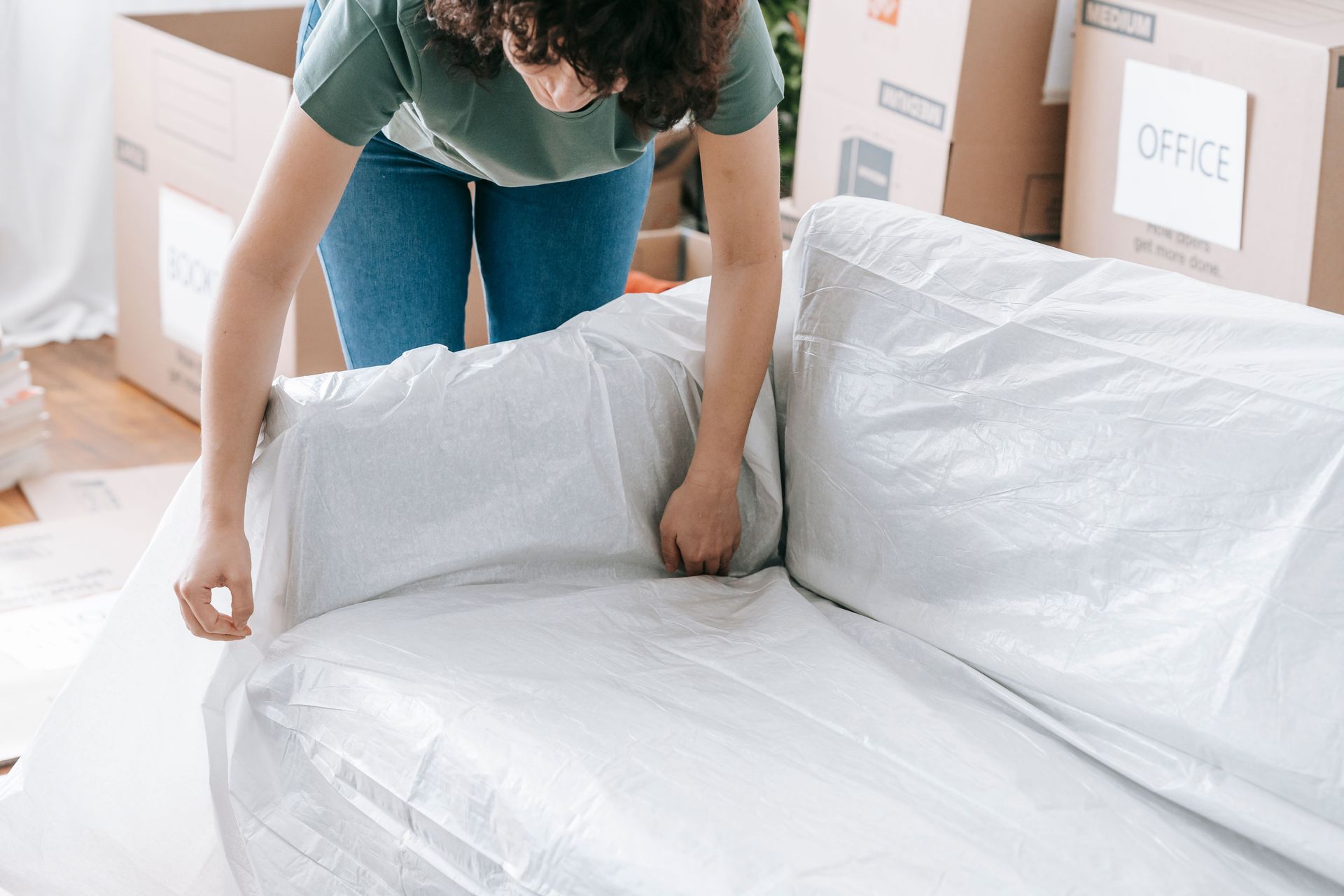 A woman carefully wraps a sofa in protective material while packing boxes in a bright room.